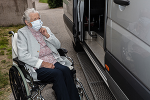 An elderly woman wearing a medical mask gets out of a disabled car