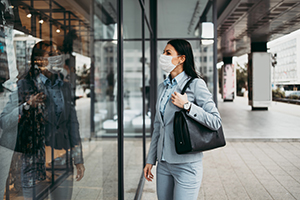 Young and elegant business woman with face protective mask standing on empty street and looking at storefront of closed store